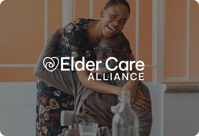 A smiling, middle-aged woman embraces an older man who is seated and also smiling. The image has the text "Elder Care Alliance" with a logo of a heart incorporated into the letter "E" overlaid on it. Both are indoors with a bright setting.
