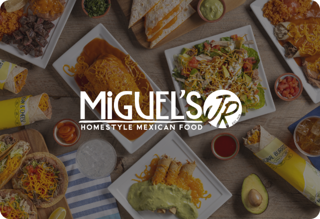 A variety of Mexican dishes are displayed on a table, including tacos, burritos, enchiladas, and a salad. Miguel's Jr. Homestyle Mexican Food logo is overlaid on the image. Side items like guacamole, salsa, and a whole avocado are also visible.