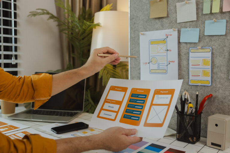 A person reviews printed wireframe designs for a mobile app at a desk, with a laptop, phone, and stationery nearby. More design sketches and sticky notes are pinned to a board in the background.