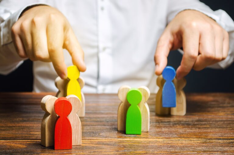 A person wearing a white shirt arranges wooden figurines on a table. The figurines are painted in different colors: red, yellow, green, and blue. The scene suggests organization or strategy.