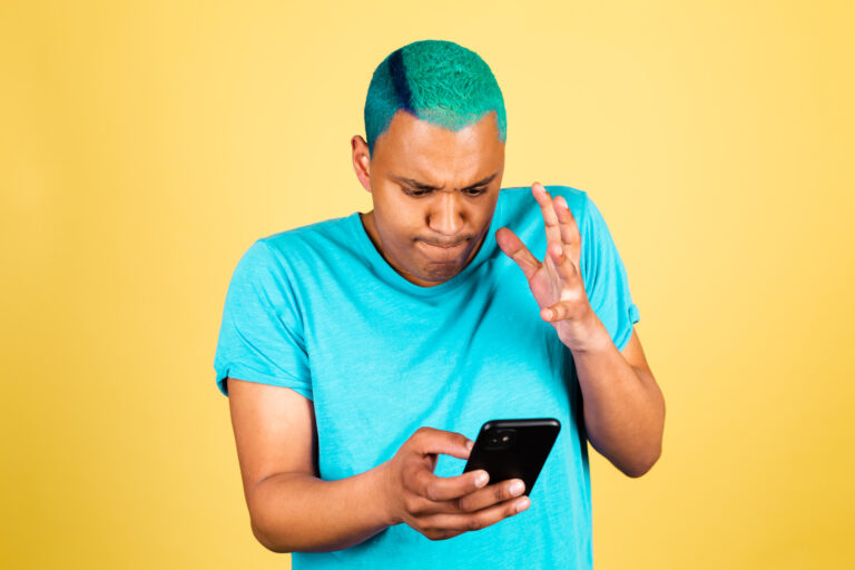 A person with short, bright blue-green hair and a matching T-shirt is looking at a smartphone with a furrowed brow and raised hand, expressing frustration or confusion. The background is solid yellow.