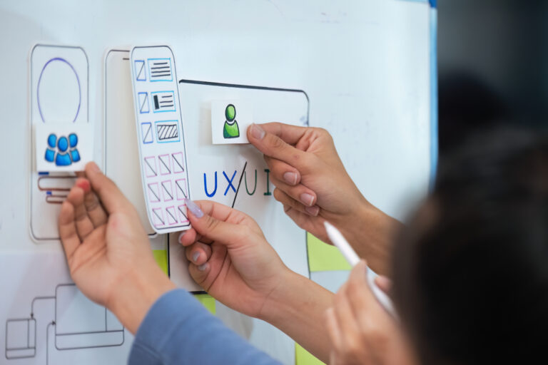 Several hands place paper cutouts of icons and interface elements onto a whiteboard with sketches and the text "UX/UI" as part of a user experience design planning session.