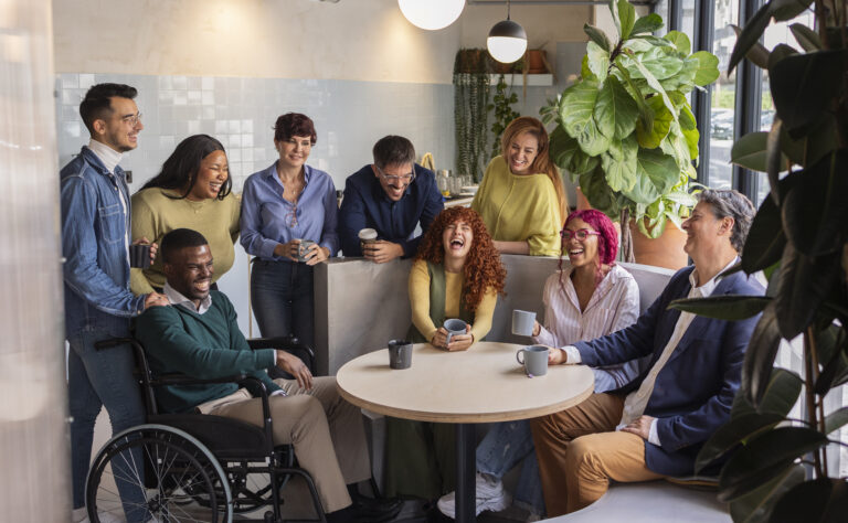 A diverse group of nine adults, including a man in a wheelchair, gather around a table with mugs, smiling and laughing in a modern, plant-filled room.
