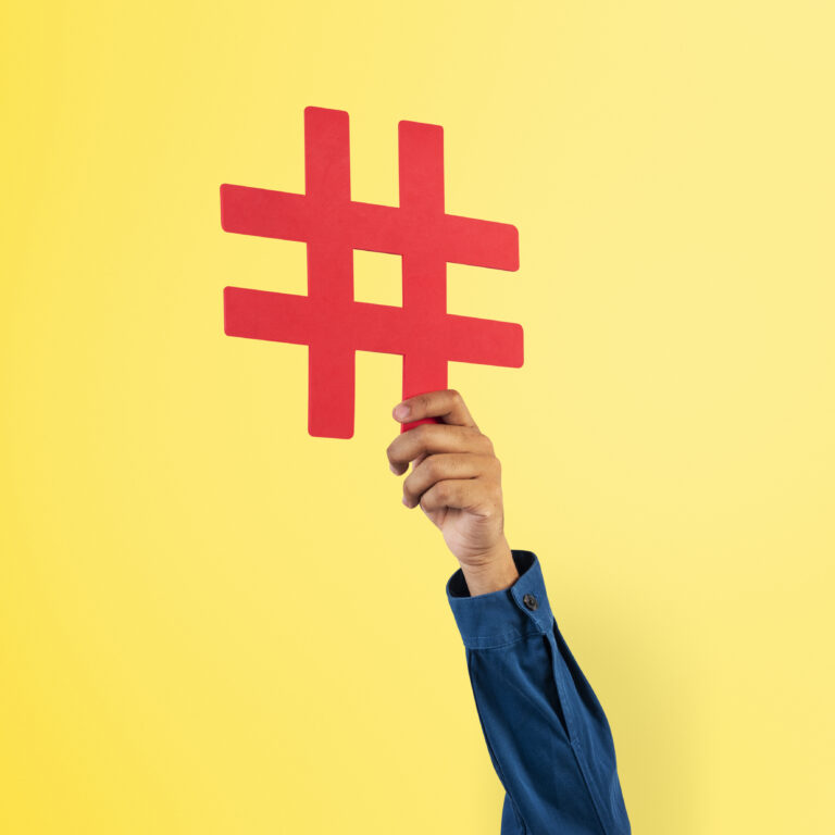 A hand in a blue sleeve is holding up a large red hashtag symbol against a yellow background.