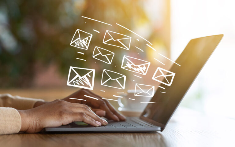 A person types on a laptop keyboard at a wooden table. Several illustrated envelope icons appear above the laptop screen, indicating sending or receiving emails. The background is softly blurred.