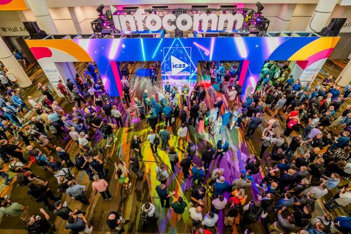 Crowd of people gathered in a brightly lit convention hall under a large "InfoComm" sign, with colorful lights and displays, viewed from above.