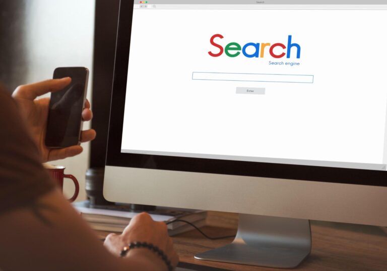 A person holding a smartphone and sitting at a desk in front of a computer monitor displaying a search engine homepage. The screen shows a search bar and the word "Search" in large, colorful letters. The desk contains a few items, including a red mug.