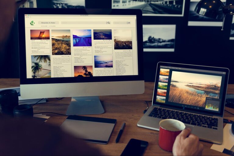 A person working at a desk with an Apple desktop computer and a laptop. The desktop screen displays a photo gallery, while the laptop shows an image editing application in landscape format. A coffee mug, smartphone, notebook, and camera lens are also visible on the desk.