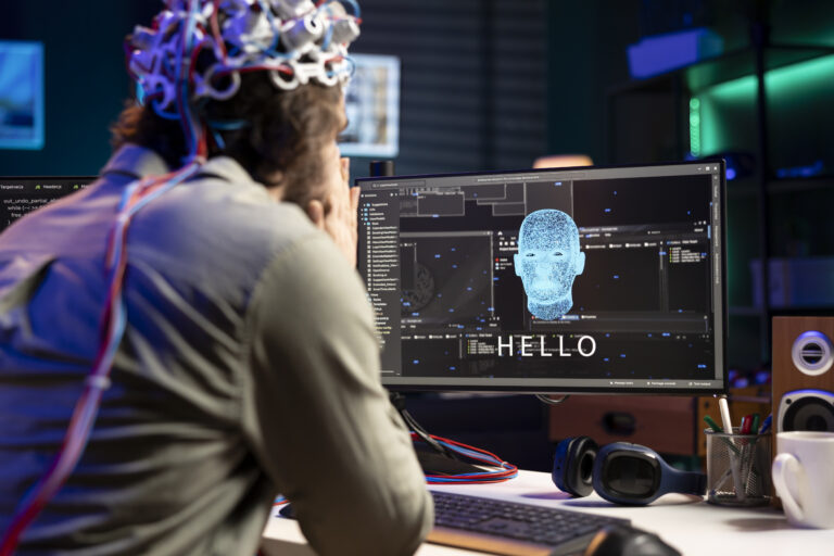 A person wearing a sensor-laden headset sits at a desk, looking at a computer screen displaying a digital face with the word "HELLO" above it. The workspace is equipped with headphones, speakers, and various tech accessories.