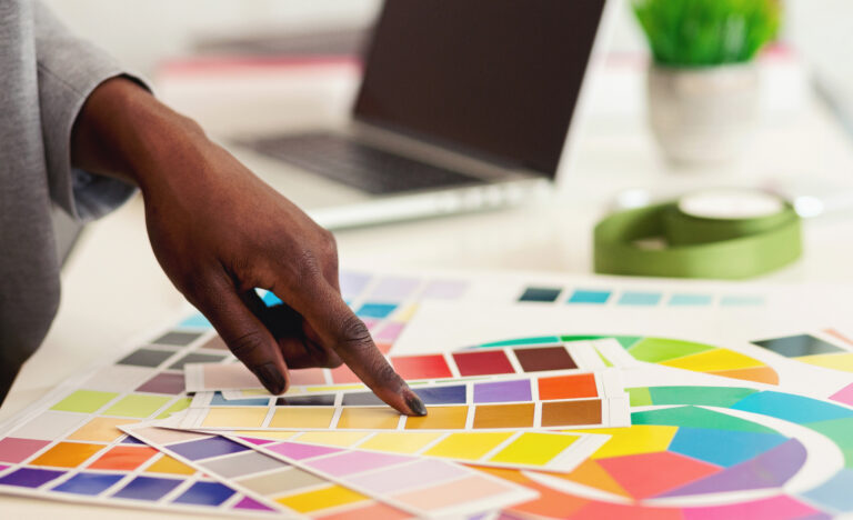 A person points at a yellow color swatch on a color palette, with various other color samples spread out on a desk. A laptop, tape, and a green plant are visible in the blurred background.