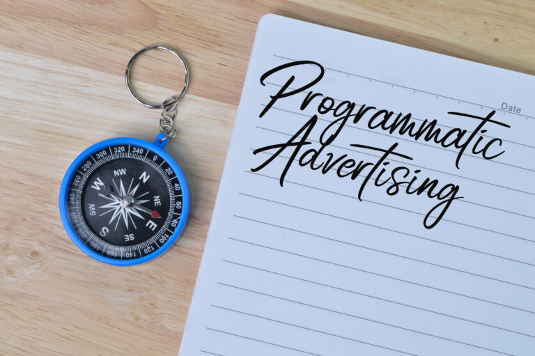 A blue compass keychain rests on a wooden surface next to an open notebook. The notebook has the words "Programmatic Advertising" written in cursive on a lined page.