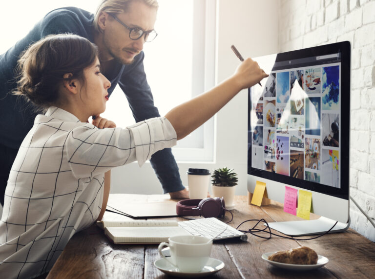 A woman sitting at a desk points at images on a large computer monitor while a man stands beside her, both focused on the screen. The desk has a keyboard, notebook, headphones, coffee cup, and sticky notes.